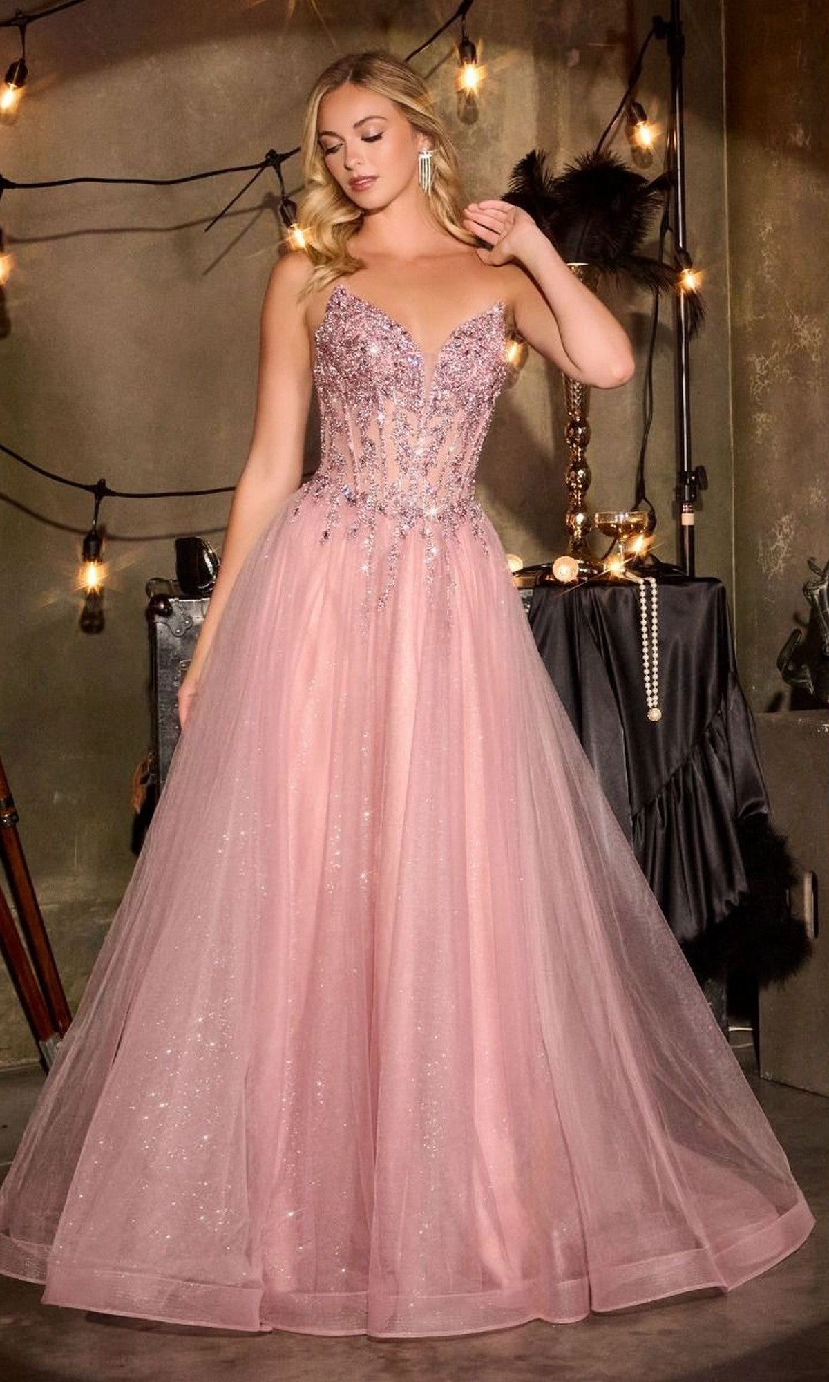 A woman wears the Lace-Up Strapless Prom Ball Gown Ladivine CD721 with a beaded corset and pink tulle skirt, standing indoors near string lights and décor.